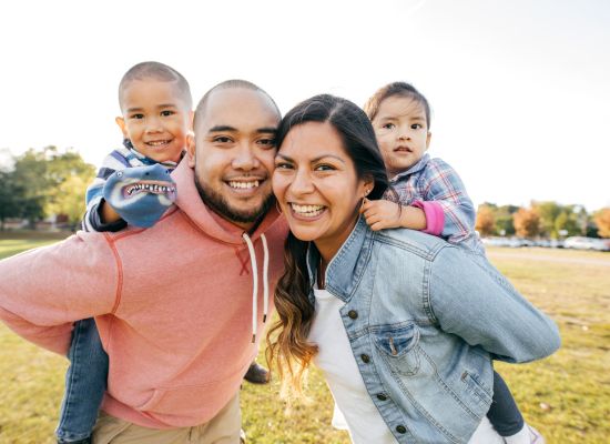 A family of four smiling at a camera standing in the park