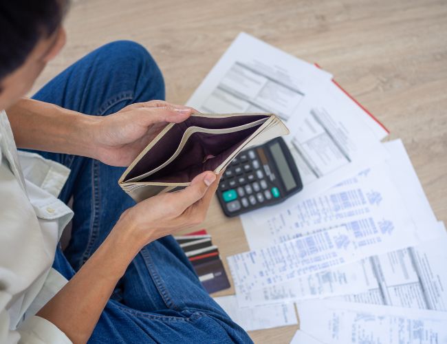 A person wearing blue jeans opening an empty wallet with a calculator and papers sitting on the floor